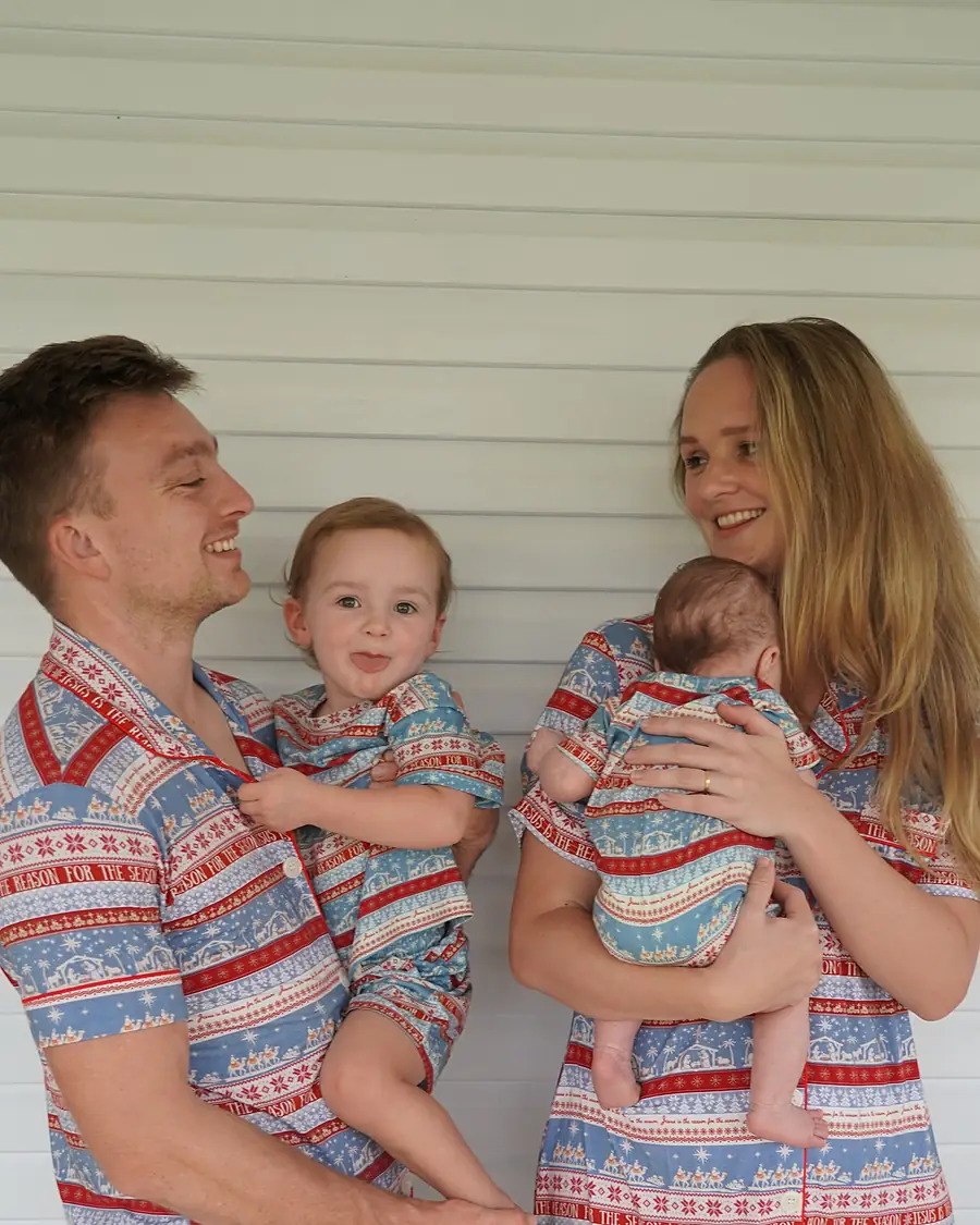 A father holding his toddler and a mother holding her baby, the family in matching O Holy Night pyjamas on a verandah